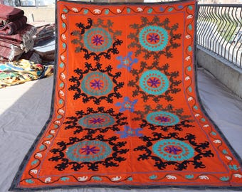 Könnte beinhalten: Ein orangefarbenes Textil mit einem sich wiederholenden Muster aus kreisförmigen Designs in Türkis, Braun und Lila. Das Textil hat einen dunklen Rand mit einem sich wiederholenden weißen Elefantenmuster. Es ist wahrscheinlich ein Teppich oder Wandbehang.
