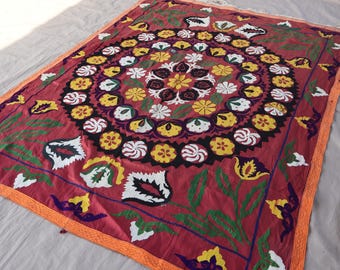 May include: A rectangular, red textile with a central circular floral design. The design features yellow, white, and purple flowers, surrounded by a border of green leaves and orange trim. The textile is likely a decorative piece.