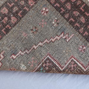 K&ouml;nnte beinhalten: Nahaufnahme eines Abschnitts eines Vintage-Teppichs mit geometrischem Muster. Der Teppich hat ein graues Mittelfeld mit Zickzack-Design und Bord&uuml;ren in Braun- und Rosat&ouml;nen. Fransen sind am Rand sichtbar.