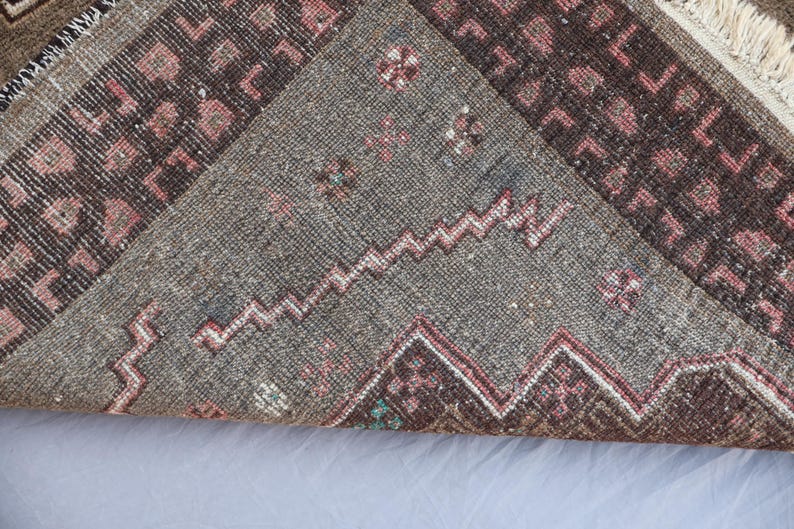 K&ouml;nnte beinhalten: Nahaufnahme eines Abschnitts eines Vintage-Teppichs mit geometrischem Muster. Der Teppich hat ein graues Mittelfeld mit Zickzack-Design und Bord&uuml;ren in Braun- und Rosat&ouml;nen. Fransen sind am Rand sichtbar.