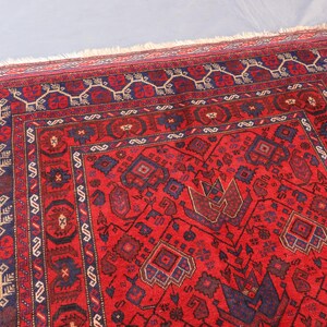 May include: A red oriental rug with intricate geometric patterns. The rug features a central field of red with dark blue and brown accents. The border is a darker red with repeating geometric designs. The rug has a fringed edge.