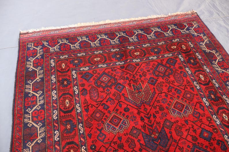May include: A red oriental rug with intricate geometric patterns. The rug features a central field of red with dark blue and brown accents. The border is a darker red with repeating geometric designs. The rug has a fringed edge.