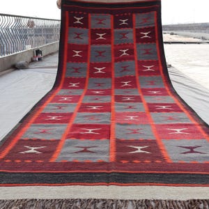 May include: A red, grey, and black patterned rug with a geometric design. The rug has a fringe border and is made of wool.