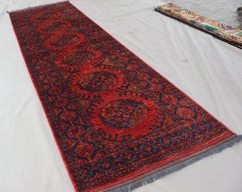 May include: A long, rectangular red rug with a detailed, repeating pattern in navy blue and touches of other colors. The rug has a fringed edge and is likely made of woven material. The design features medallions and geometric shapes.