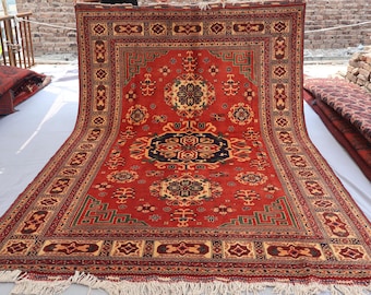May include: A large, rectangular red rug with an intricate geometric pattern. The rug features a central medallion in black and navy blue, surrounded by floral and geometric motifs in beige, brown, and green. The rug has a fringed edge.