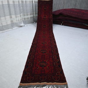 May include: A long, rectangular, red rug with a detailed geometric pattern. The rug has a dark red background with black and gold accents. The edges are finished with a fringe. Several other rugs are stacked in the background.
