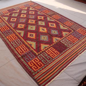 May include: A rectangular, hand-woven rug with a geometric pattern. The rug features a deep red background with repeating diamond shapes in yellow, green, and blue. The border has a repeating geometric design in orange and purple. The rug is approximately 6 feet by 9 feet.