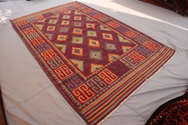 May include: A rectangular, hand-woven rug with a geometric pattern. The rug features a deep red background with repeating diamond shapes in yellow, green, and blue. The border has a repeating geometric design in orange and purple. The rug is approximately 6 feet by 9 feet.