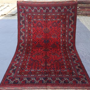 May include: A rectangular, hand-knotted rug with a deep red field and intricate geometric patterns in navy blue and cream. The rug features a central medallion design and a detailed border. The rug is displayed on a light gray surface.