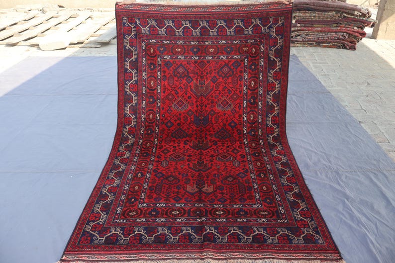 May include: A rectangular, hand-knotted rug with a deep red field and intricate geometric patterns in navy blue and cream. The rug features a central medallion design and a detailed border. The rug is displayed on a light gray surface.