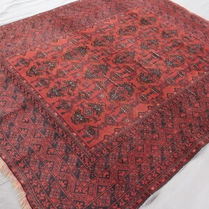 Pode incluir: Um tapete oriental vermelho com um padr&atilde;o geom&eacute;trico detalhado em preto. O tapete apresenta um design central rodeado por uma borda com formas repetidas e franjas nas bordas. O tapete &eacute; provavelmente feito de l&atilde;.