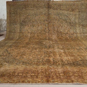 May include: A large, rectangular, vintage rug with an intricate floral pattern in shades of brown, gold, and green. The rug has a distressed appearance, suggesting age and use. The edges are slightly frayed, adding to its antique charm. The rug is displayed on a neutral surface.