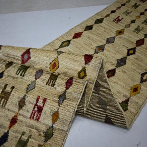 May include: A long, rectangular runner rug with a beige, textured base. The surface features repeating vertical rows of colorful, diamond-shaped geometric patterns and stylized animal figures in shades of red, green, yellow, and dark blue.