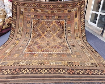May include: A large rectangular area rug featuring a central diamond pattern in shades of brown, tan, and cream. The border displays intricate geometric motifs, including small crosses and stylized symbols in muted orange, yellow, and dark brown tones.