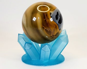 May include: A golden sphere with brown and black swirls sits on a blue crystal stand. The stand has a circular base and multiple crystal points.