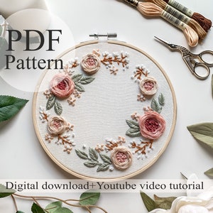 May include: A PDF pattern for a floral embroidery hoop art project. The hoop features a wreath of pink roses, green leaves, and white flowers. The image also includes embroidery floss, scissors, and a candle.