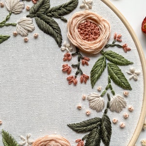 May include: A close-up of a hand-stitched embroidery hoop with a floral design. The design features peach and white flowers with green leaves, all stitched on a white fabric.
