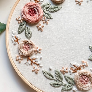 May include: A circular embroidery hoop with a white fabric background featuring a floral design. The design includes pink and beige roses, green leaves, and small white flowers.