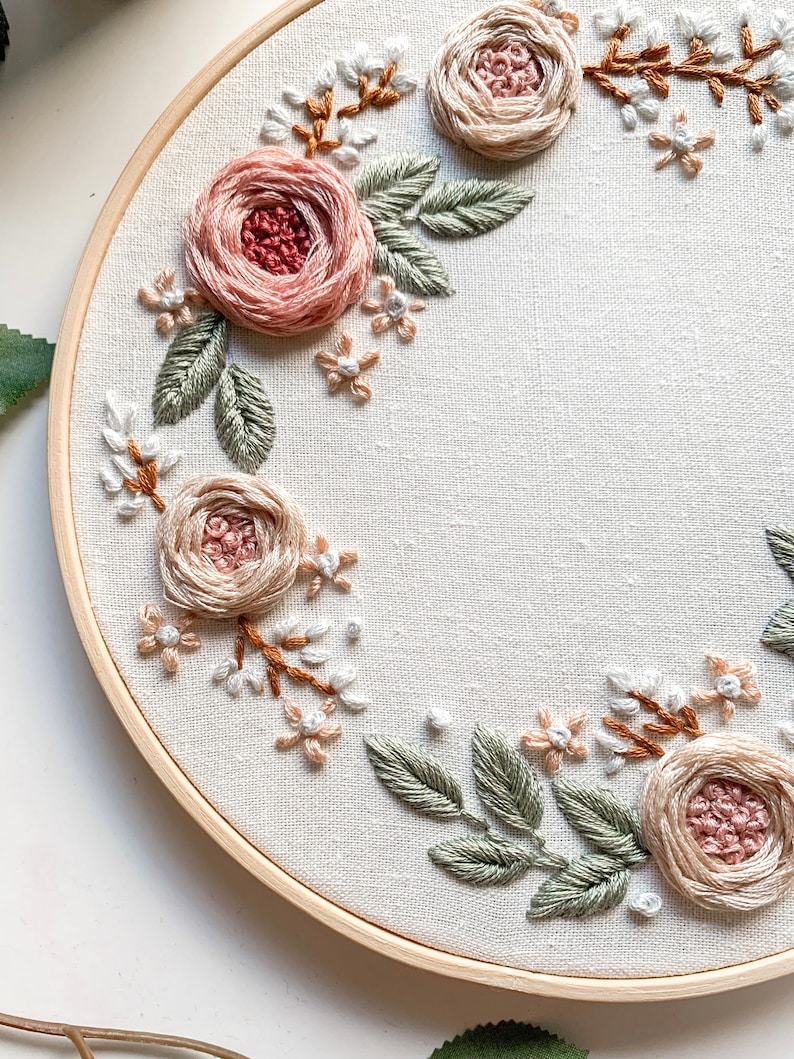 May include: A circular embroidery hoop with a white fabric background featuring a floral design. The design includes pink and beige roses, green leaves, and small white flowers.