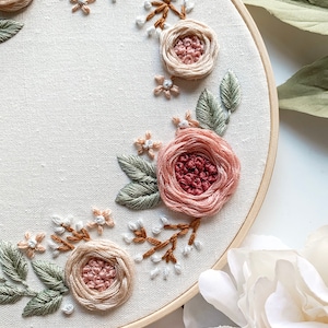May include: A close-up of a circular embroidery hoop with a floral design. The design features pink and red roses, green leaves, and white and brown stems. The embroidery is on a white fabric background.