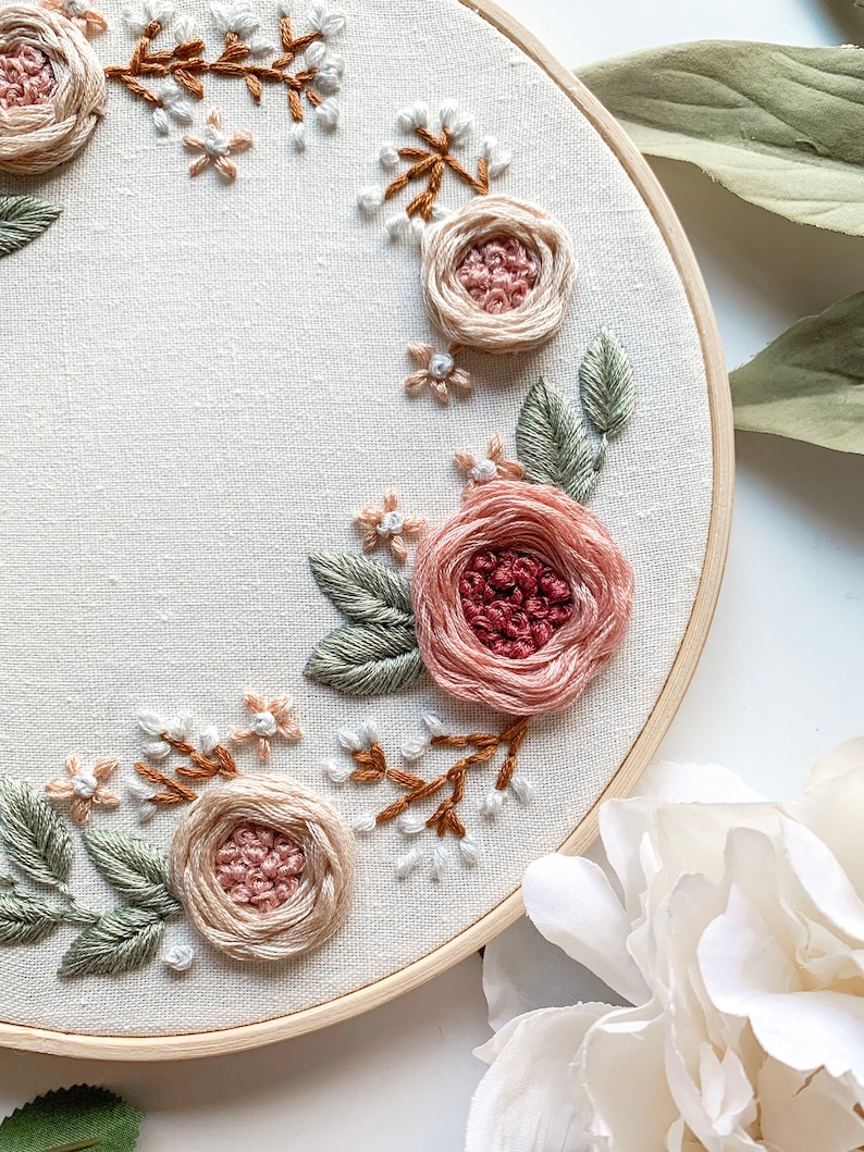 May include: A close-up of a circular embroidery hoop with a floral design. The design features pink and red roses, green leaves, and white and brown stems. The embroidery is on a white fabric background.