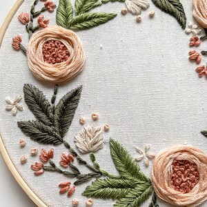May include: A close-up of a floral embroidery hoop art piece. The hoop is made of wood and has a white fabric background. The embroidery features a wreath of peach and white flowers with green leaves.