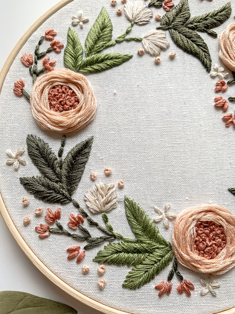 May include: A close-up of a floral embroidery hoop art piece. The hoop is made of wood and has a white fabric background. The embroidery features a wreath of peach and white flowers with green leaves.
