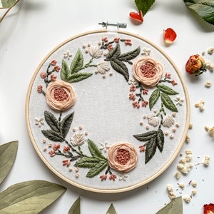 May include: Embroidered hoop art featuring a wreath of peach-colored flowers and green leaves on a white background.