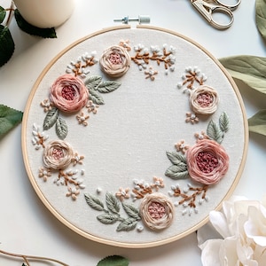 May include: A circular embroidery hoop with a floral wreath design. The wreath is made of pink and beige roses, green leaves, and white flowers. The hoop is made of wood and has a natural finish.