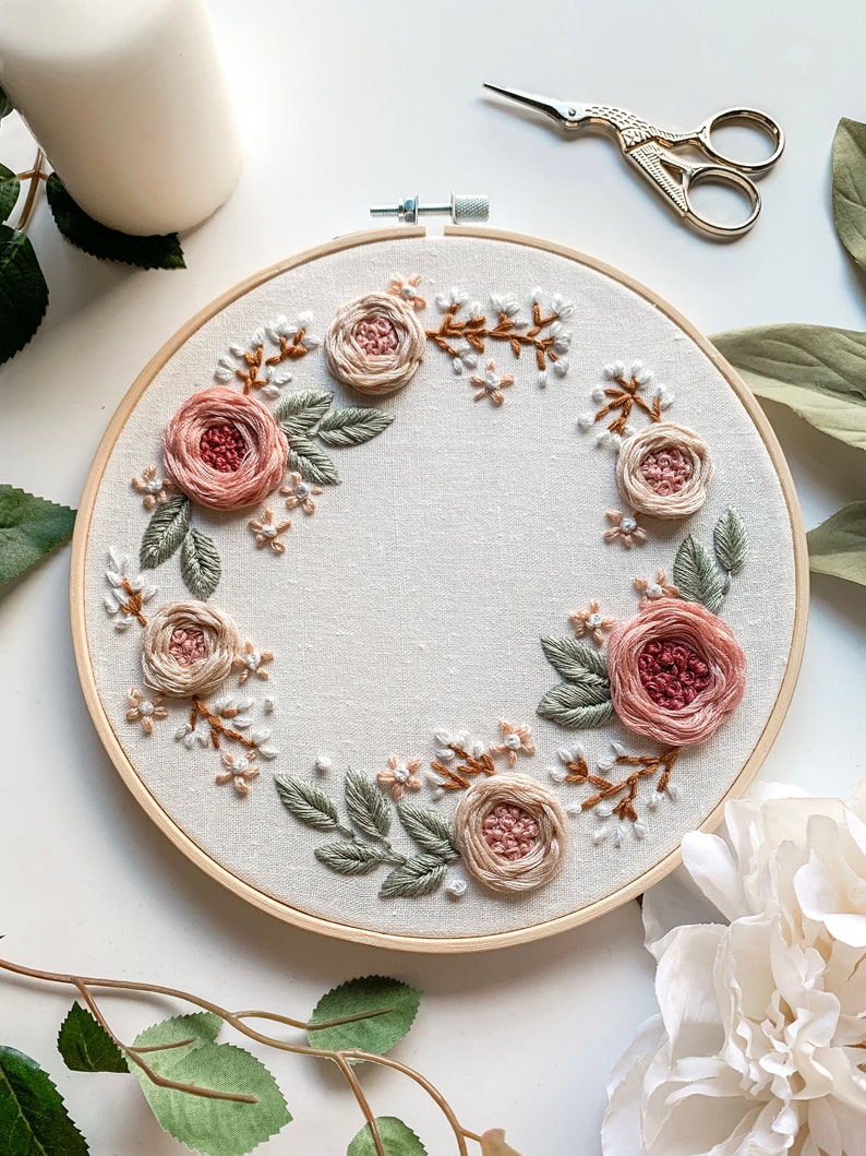 May include: A circular embroidery hoop with a floral wreath design. The wreath is made of pink and beige roses, green leaves, and white flowers. The hoop is made of wood and has a natural finish.