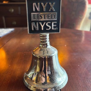 May include: A silver bell with a black rectangular top. The top has the text "NYX LISTED NYSE" in silver lettering. The bell is sitting on a dark wooden surface.