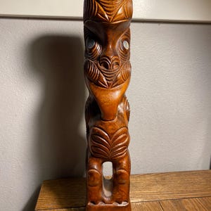 May include: A carved wooden tiki statue with intricate details. The brown statue features a stylized face with inlaid eyes and a carved body. The tiki is standing upright and has a rectangular base. The statue is on a wooden surface.