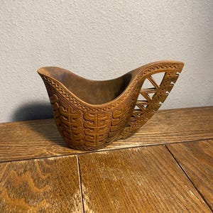 May include: A carved wooden bowl with a handle. The bowl is brown and features intricate geometric patterns and designs. The handle has a unique openwork design. The bowl is sitting on a wooden surface.