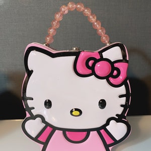 May include: Pink Hello Kitty lunchbox with a pink and black handle. The lunchbox features a white Hello Kitty face with a pink bow and pink dress.