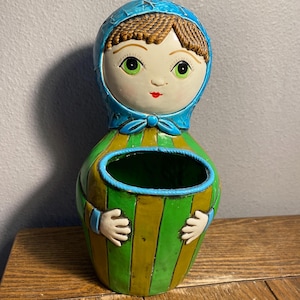 May include: A ceramic planter in the shape of a doll. The doll has a blue bonnet, green eyes, and a red mouth. The body is green and yellow striped with a blue rim and small white hands. The planter is on a wooden surface.