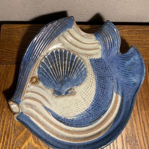 May include: A ceramic fish-shaped serving dish with a blue and white glaze. The dish features a seashell design in the center and wave-like patterns. The edges are textured, and the dish is placed on a wooden surface.
