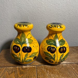 May include: A pair of yellow ceramic salt and pepper shakers. Each shaker has a bulbous body with a narrow neck and a rounded top. The shakers are decorated with a cherry design and the words "PEPE" and "SALE" are written on top.
