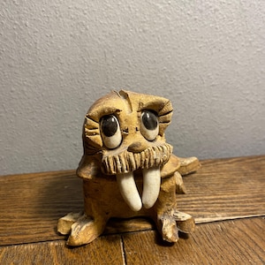 May include: A ceramic figurine of a walrus with large eyes, a brown body, and white tusks. The figurine is sitting on a wooden surface.