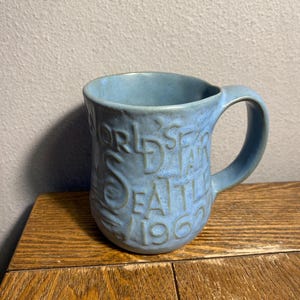 May include: A light blue ceramic mug with a handle. The mug has embossed text that reads "WORLD'S FAIR SEATTLE 1962". The mug is sitting on a wooden surface.