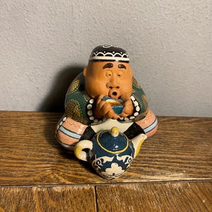 May include: A ceramic figurine of a seated person drinking from a small bowl. The figure wears a patterned hat and robe, with a teapot in front. The teapot is blue with white and yellow accents. The figurine is on a wooden surface.