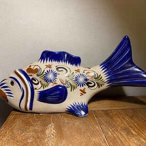 May include: A ceramic fish figurine with a cream-colored body, adorned with blue, brown, and green floral patterns. The fish has blue fins and tail, and is resting on a wooden surface. This decorative item is suitable for home decor.