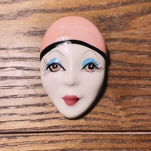 May include: A ceramic mask with a pink headband, white face, black eyelashes, blue eyeshadow, and a pink mouth.