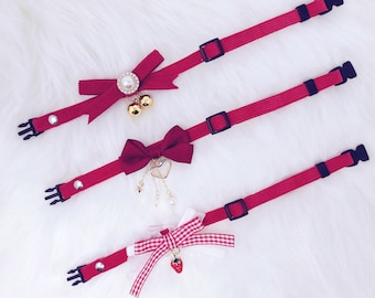 May include: Three red cat collars with different charms. The first collar has a red bow with a pearl and gold bells. The second collar has a red bow with a gold heart and dangling charms. The third collar has a red and white gingham bow with a strawberry charm.