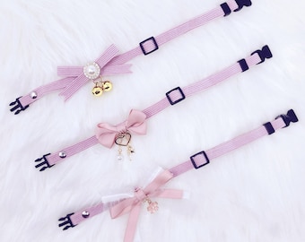May include: Three pink cat collars with different charms. The first collar has a pink bow with a pearl and two bells. The second collar has a pink bow with a heart charm and a pearl. The third collar has a pink bow with a flower charm.