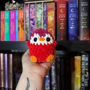 May include: A handmade, plush penguin toy in red, white, orange, and burgundy. The penguin has black eyes and a white belly. The toy is crocheted and has orange feet. The background includes a bookshelf with colourful books.