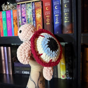 May include: A handmade crochet turtle plushie with a unique design. The turtle's body is light pink, and the shell is designed to look like an eye, with a red border, a black pupil, and blue and white details. The plushie is soft and cuddly.