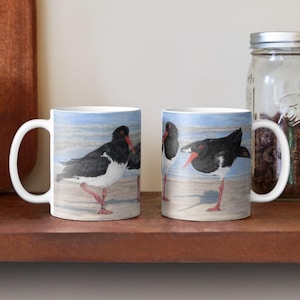 May include: Two white ceramic mugs featuring a painted image of oyster catchers on a beach. The birds are black and white with red beaks and legs. The background shows a light blue sky and sand.