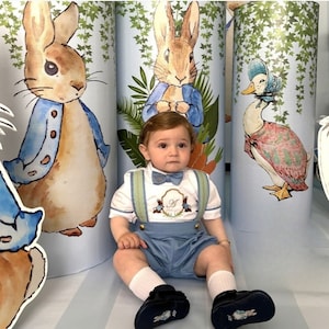 Peut inclure: Un bébé garçon portant une salopette bleu clair avec des bretelles et un nœud papillon. La salopette est ornée d'un motif brodé de deux lapins et de la lettre "H". Le bébé est assis devant une toile de fond avec des illustrations de personnages de Peter Rabbit.