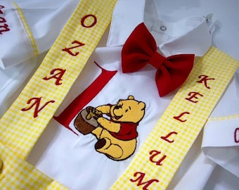 May include: A white onesie with yellow and white checked braces. The onesie has an embroidered Winnie the Pooh character and the name "OZAN KELLUM" on the braces.