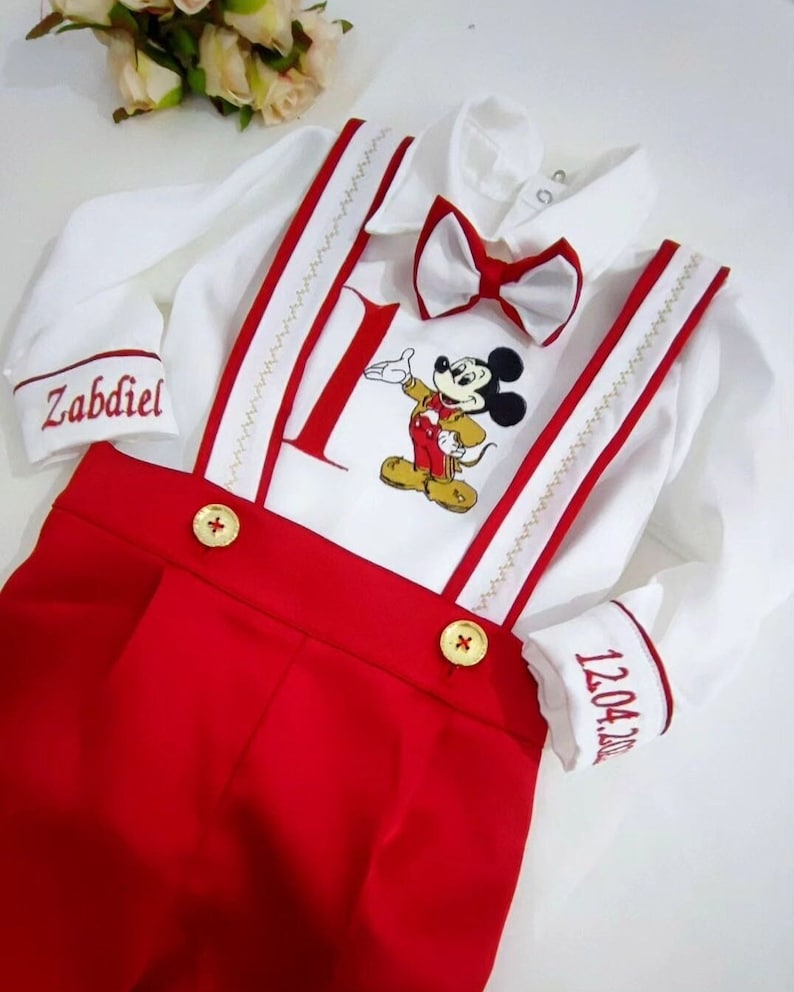 May include: A white onesie with red trim and a red bow tie. The onesie has a Mickey Mouse character embroidered on the front, along with the number "1". The onesie also has the name "Zabdiel" embroidered on one sleeve and the date "12.04.2021" embroidered on the other sleeve.